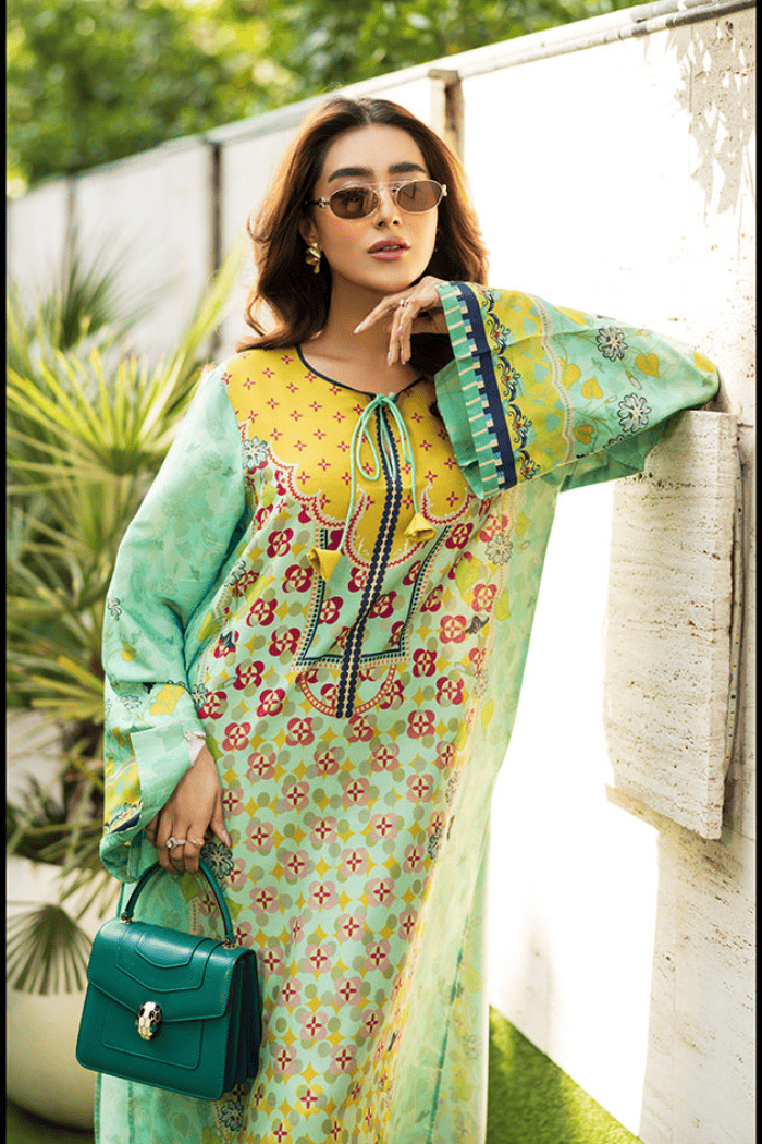Woman in a colorful dress with a green handbag standing against a white wall with plants in the background