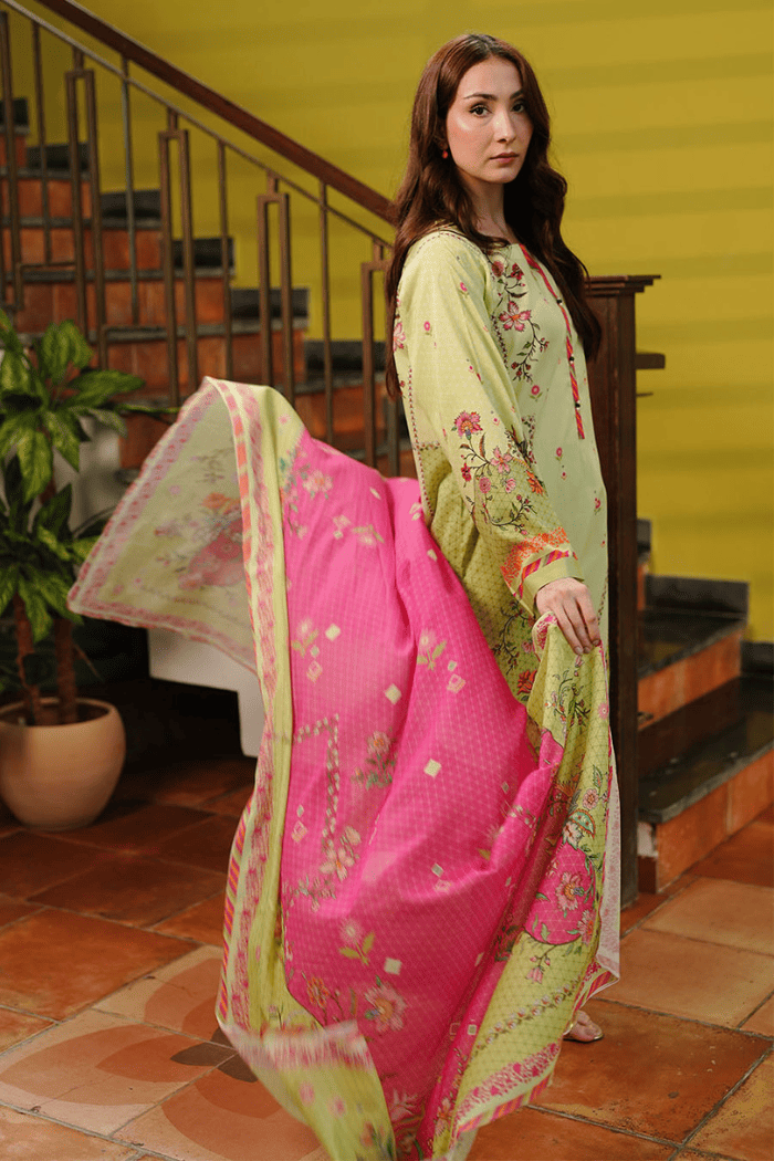 Woman in a green and pink traditional outfit standing indoors with a yellow wall and staircase in the background.