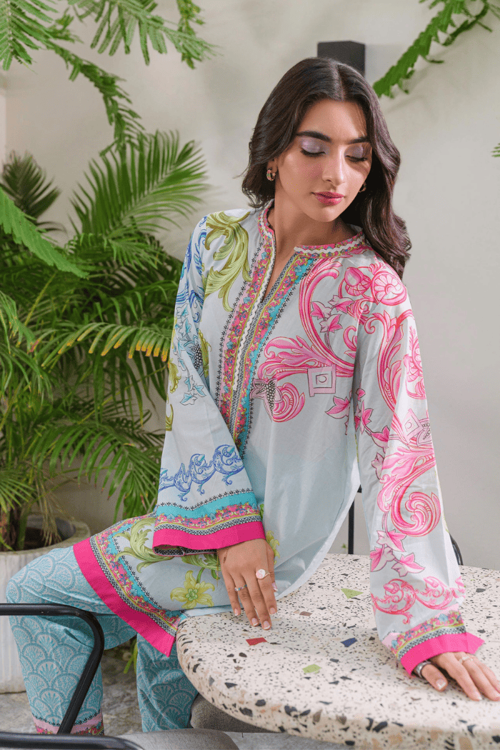 Woman wearing a light blue and pink embroidered outfit sitting on a chair with plants in the background