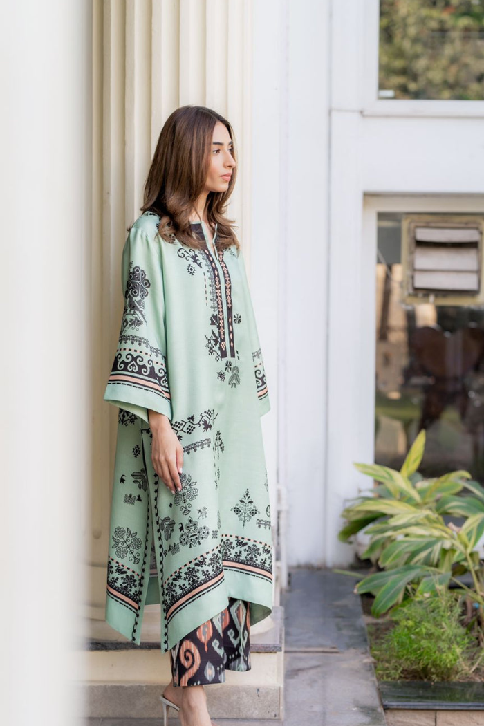 Woman wearing a light green dress with black floral patterns standing in front of a white door.