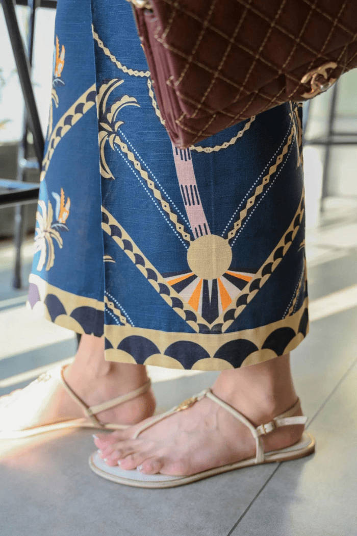 Person wearing a blue patterned skirt with gold accents, holding a brown handbag, and wearing beige sandals.