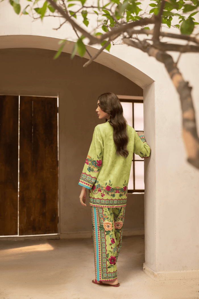Woman in a green traditional outfit with floral patterns standing in a sunlit room.