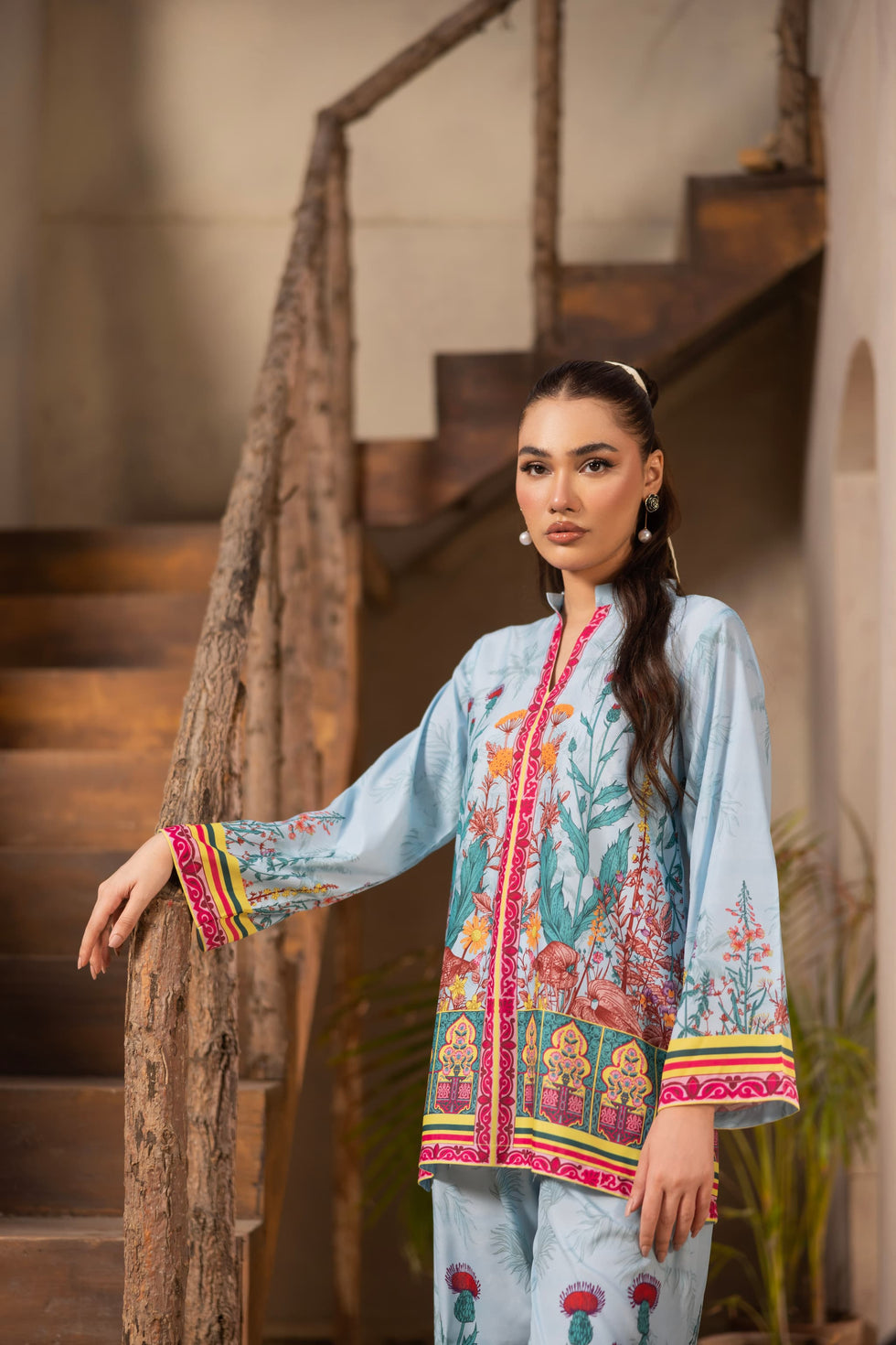 Swiss cotton sky blue co-ord set with pink, yellow, and green floral design. A woman wearing a colorful traditional outfit sis tanding in front of wooden stairs.