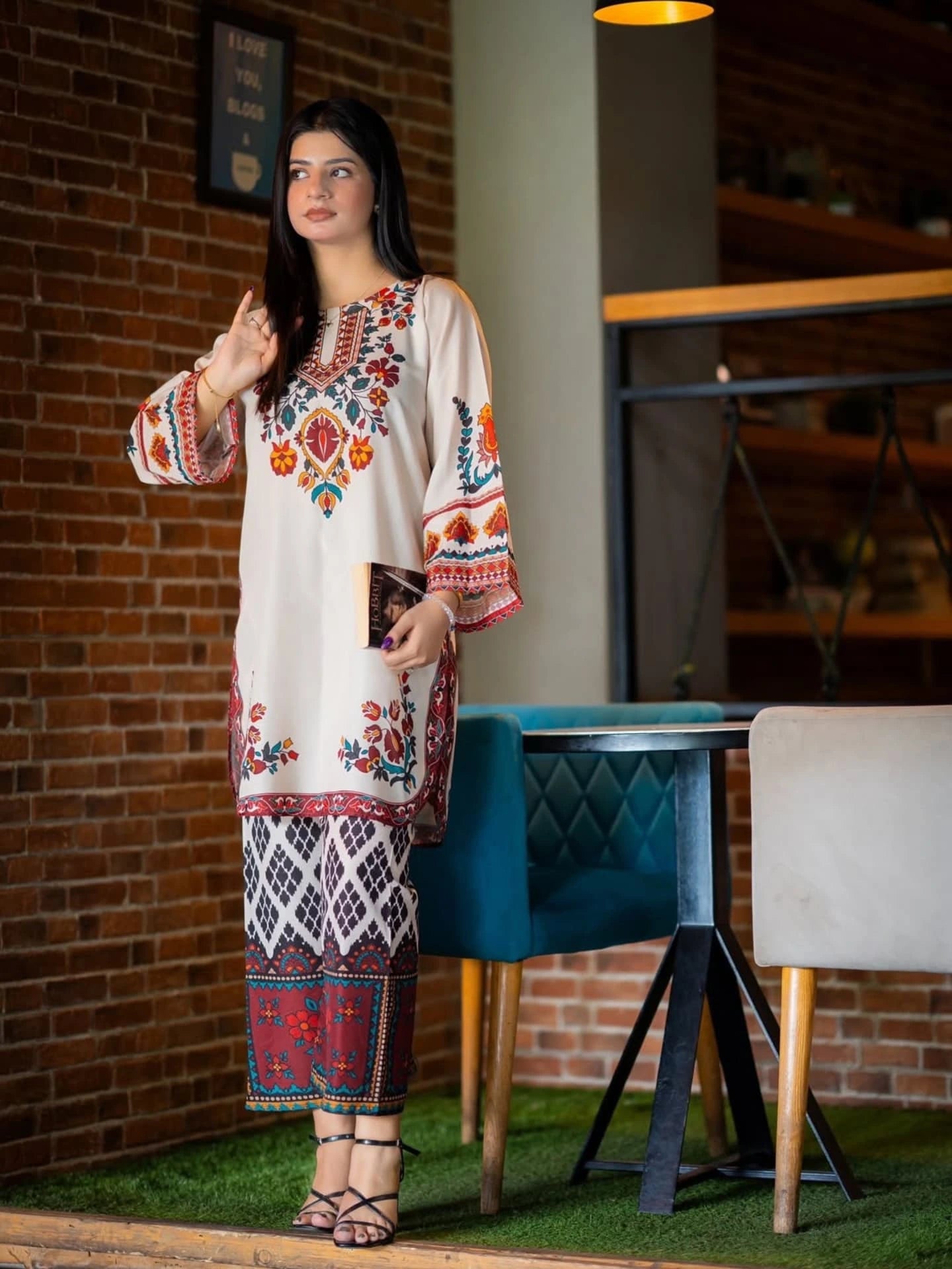 Woman in a patterned outfit standing in a casual indoor setting with a brick wall and modern furniture.